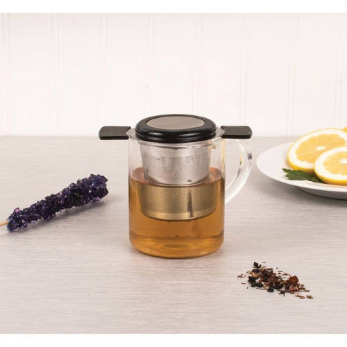 A clear glass teapot with a black lid and a metal infuser inside, filled with amber tea, set on a light tabletop with lavender, loose leaves, and lemon slices in the background.