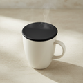 A white ceramic mug with a black lid, shown with steam rising from the top on a warm beige surface.