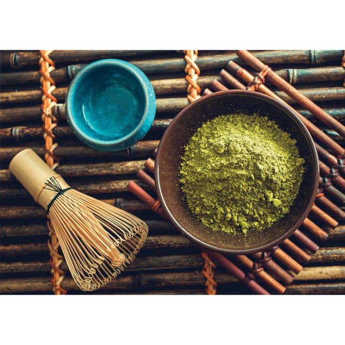 Matcha green tea powder in a bowl with a whisk and small cup on a woven mat.
