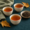 Three ivory cups of amber tea with loose tea leaves and golden autumn leaves on a teal surface.